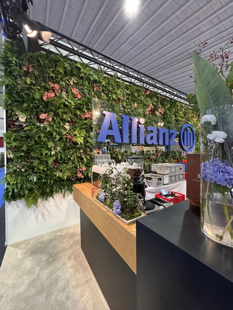 Allianz Logo vor einer Wand aus lebenden Pflanzen und einem Messestand mit Ausstellungsstücken.