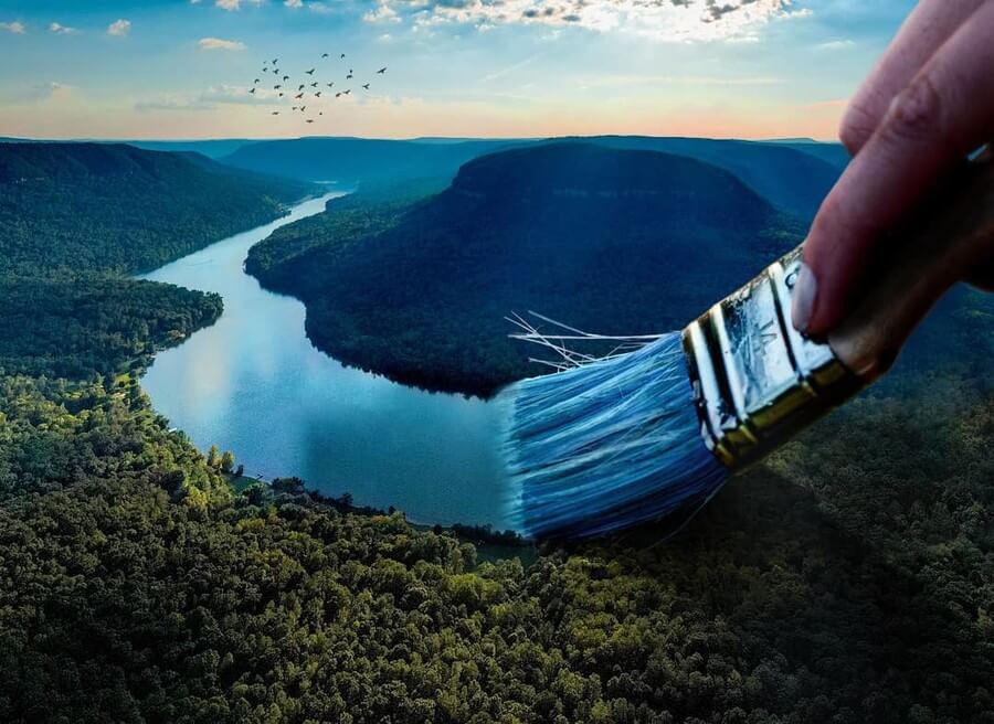 Eine Hand malt mit einem Pinsel eine Flusslandschaft in grüner Hügellandschaft unter blauem Himmel.
